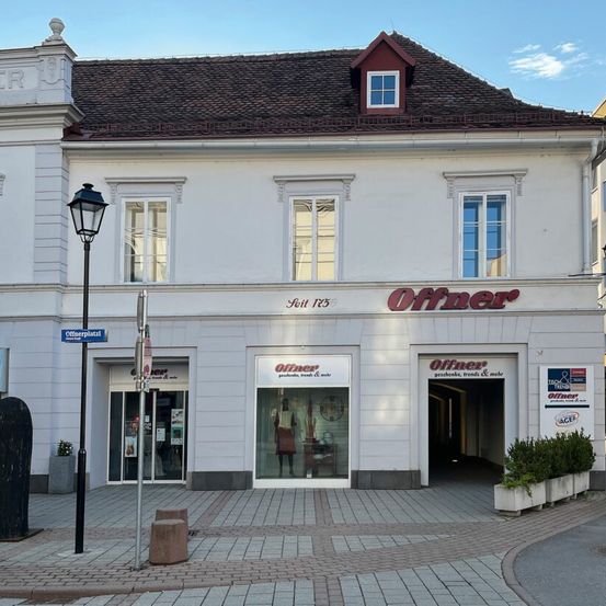 Ein weißes Gebäude mit braunem Dach und rotem Rand hat ein Schild mit der Aufschrift 'Offner'. Das Gebäude hat mehrere Fenster und eine Glastür mit einer Puppe im Inneren. Eine Straßenlampe und ein Schild mit der Aufschrift 'Offnerplatz' befinden sich auf der linken Seite.