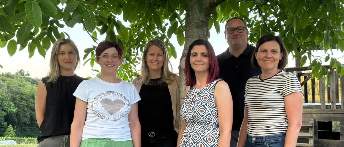 Eine Gruppe von sechs Personen, vier Frauen und zwei Männer, stehen unter einem Baum in einem Park. Sie lächeln und scheinen für ein Foto zu posieren.