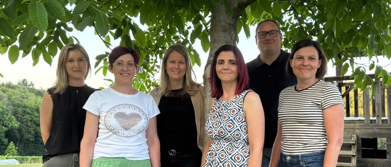 Eine Gruppe von sechs Personen, vier Frauen und zwei Männer, stehen unter einem Baum in einem Park. Sie lächeln und scheinen für ein Foto zu posieren.