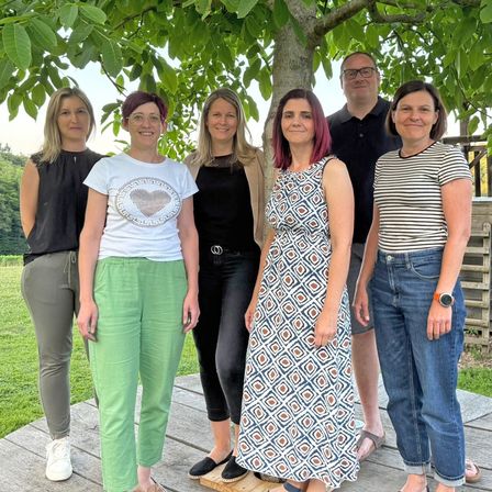 Sechs Personen stehen unter einem Baum in einem Park und lächeln für ein Foto. Sie sind lässig gekleidet und scheinen für ein Gruppenfoto zu posieren.