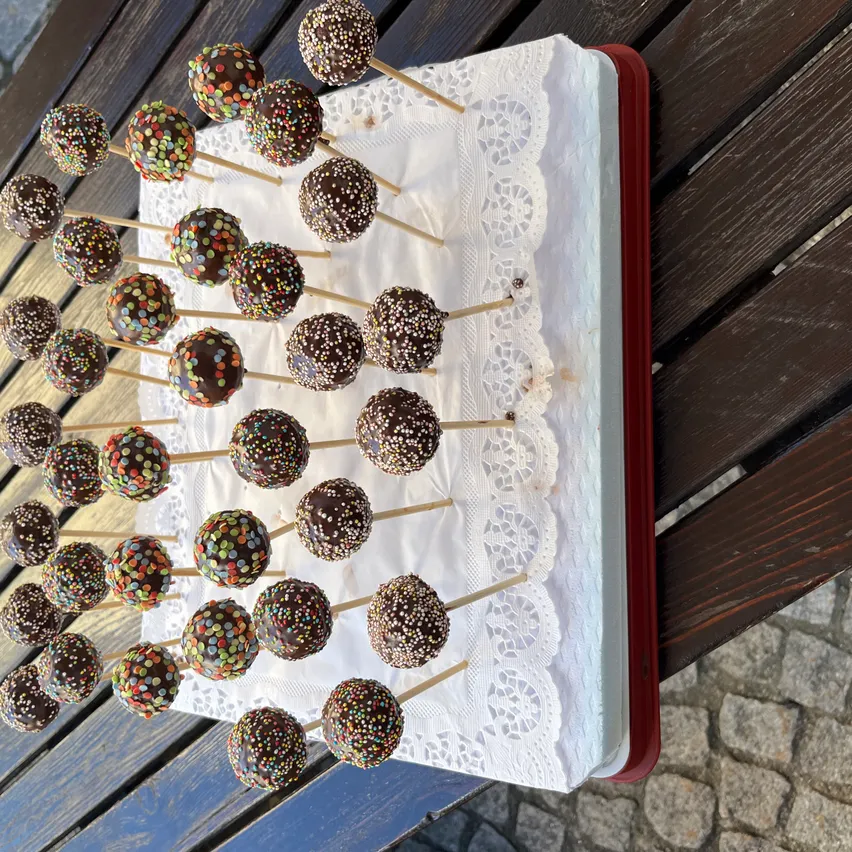 Ein Tablett mit Schokoladen-Kuchen-Pops mit bunten Streuseln steht auf einer Holzoberfläche. Die Pops sind auf einem weißen Spitzentuch und werden von Holzstäbchen gehalten.