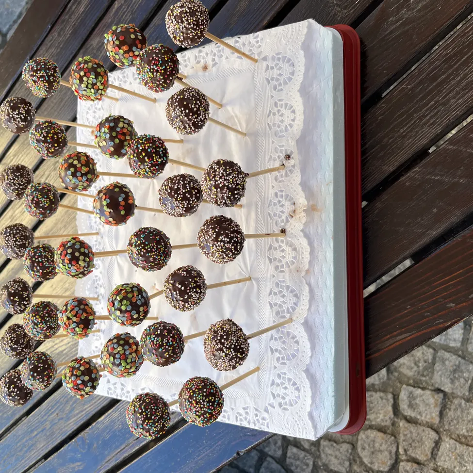 Ein Tablett mit Schokoladen-Kuchen-Pops mit bunten Streuseln steht auf einer Holzoberfläche. Die Pops sind auf einem weißen Spitzentuch und werden von Holzstäbchen gehalten.