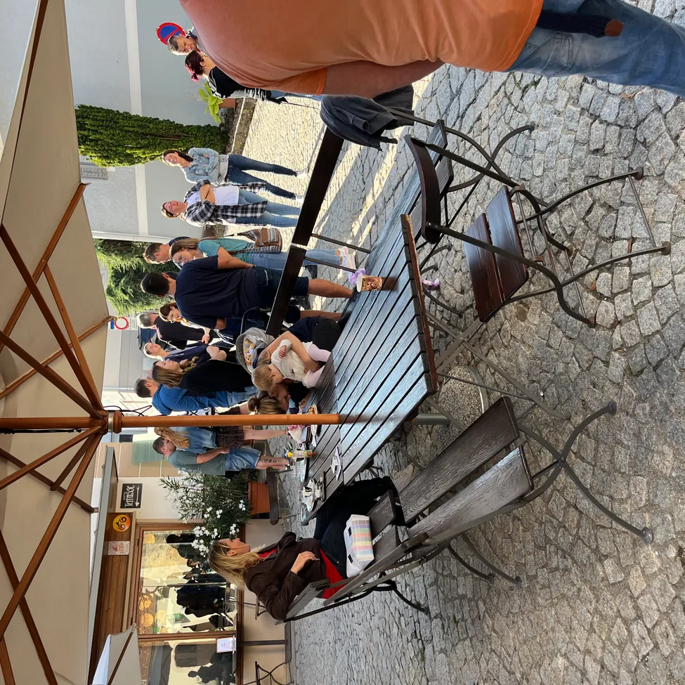 Die Aufsicht auf einen Außenbereich zeigt mehrere Menschen, die sich um einen Tisch unter einem großen Schirm versammelt haben. Einige sitzen, während andere stehen und interagieren. Der Tisch ist mit verschiedenen Gegenständen bestückt, und es gibt Pflanzen und ein Gebäude im Hintergrund.