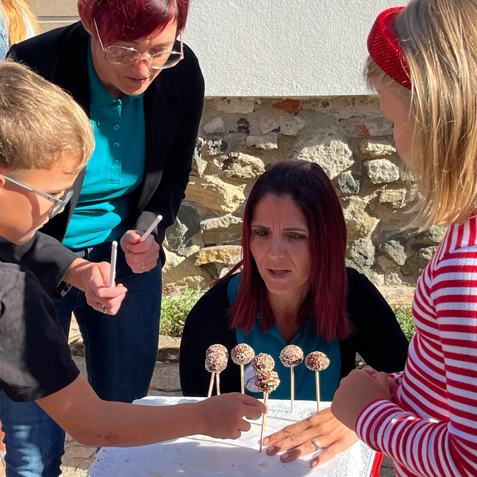 Eine Gruppe von Kindern und Erwachsenen dekoriert Kuchen-Pops bei einer Veranstaltung im Freien. Eine Frau hält einen Stock mit Kuchen-Pops, während andere zusehen.