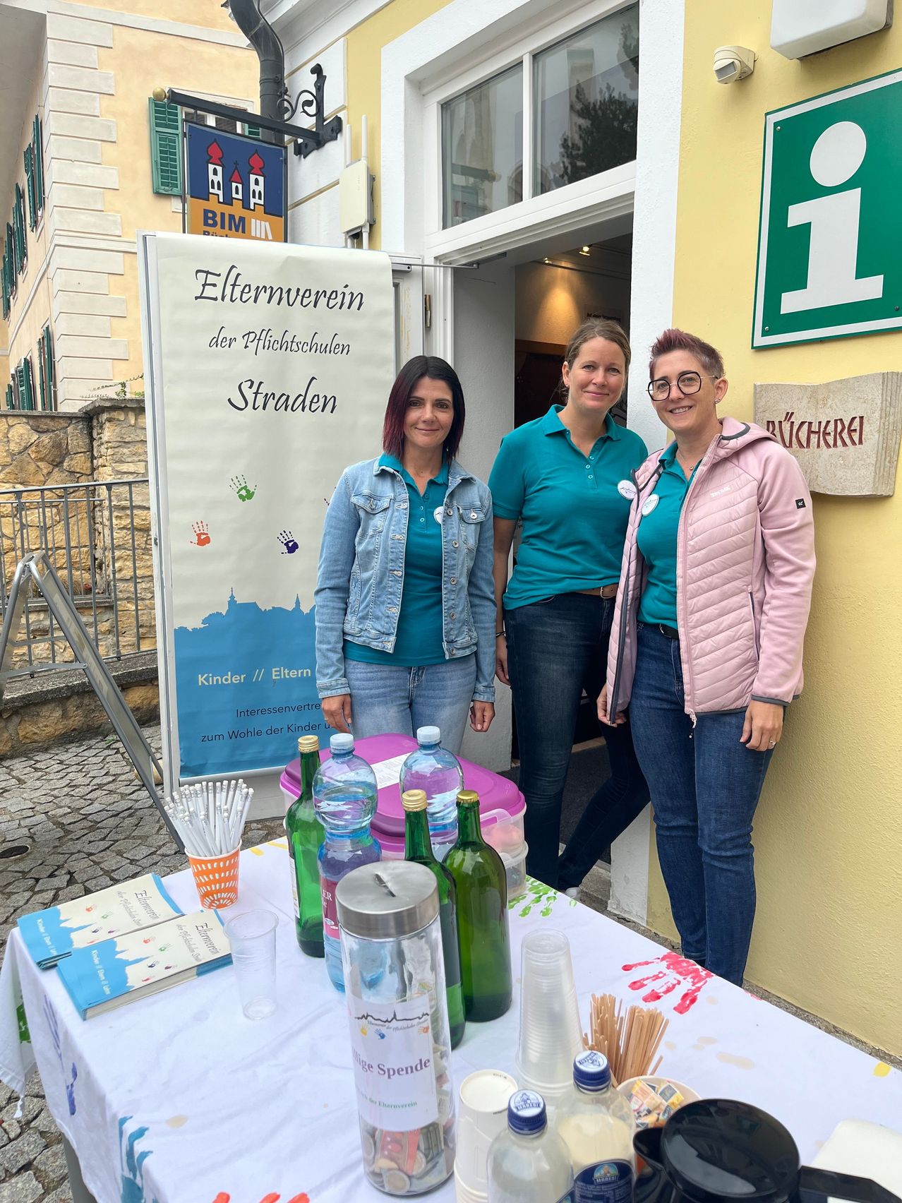 Three women stand behind a table with bottles and books, in front of a building with a sign reading 'Elternverein der Pflichtschulen Straden'.