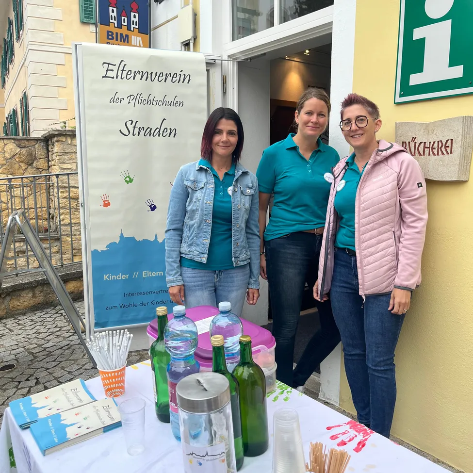 Drei Frauen stehen hinter einem Tisch mit Flaschen und Büchern vor einem Gebäude mit einem Schild, das 'Elternverein der Pflichtschulen Straden' besagt.