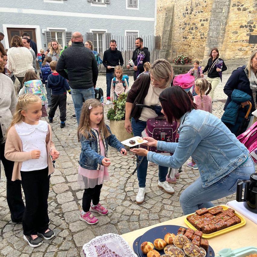 A group of adults and children are gathered around a table with food, possibly at a community event. A woman hands a plate to a young girl.