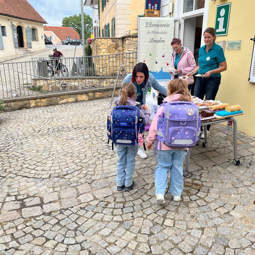Two women serve food to two young girls with backpacks in front of a building with a sign that reads 'Elternverein der Grundschulen Straden'.