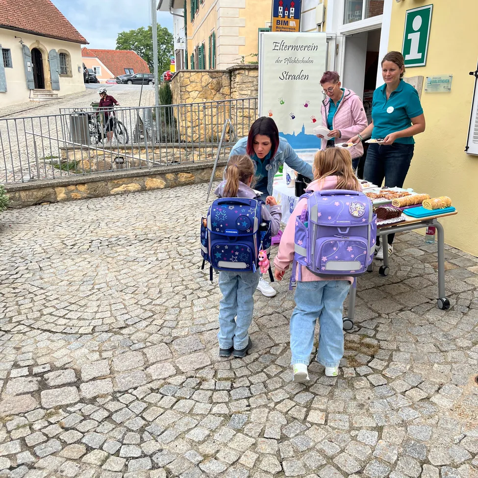 Zwei Frauen servieren Essen für zwei junge Mädchen mit Rucksäcken vor einem Gebäude mit einem Schild, das 'Elternverein der Grundschulen Straden' besagt.