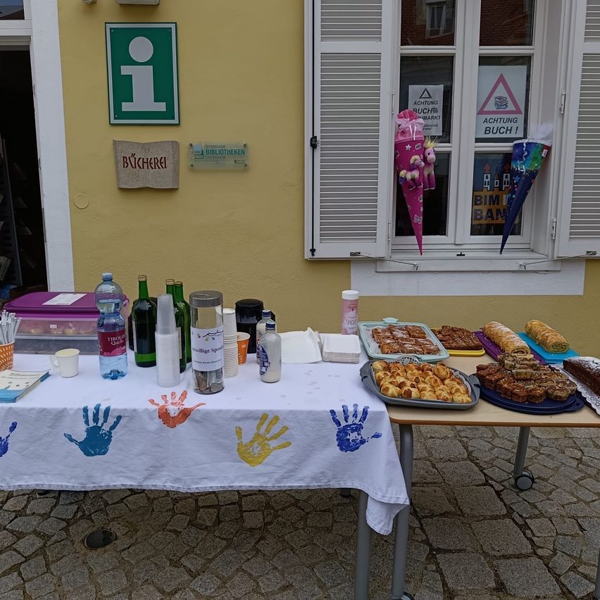A table with various food items, drinks, and handprints on a white tablecloth. A sign on the wall reads 'Bücherei'.
