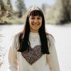 Eine Frau mit langen Haaren steht am See, hält ein Buch in der Hand, lächelt und trägt einen weißen Pullover mit Herzmuster.