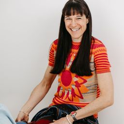 Eine Frau mit langen schwarzen Haaren lächelt und trägt ein farbenfrohes gestreiftes Shirt mit Sonnendesign. Sie sitzt auf einer weißen Oberfläche und trägt eine Armband und eine Uhr.