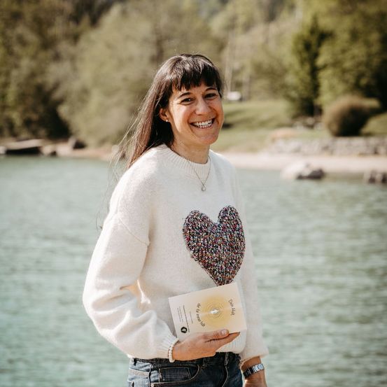 Eine Frau lächelt und hält ein Buch am See mit einem bewaldeten Ufer im Hintergrund.
