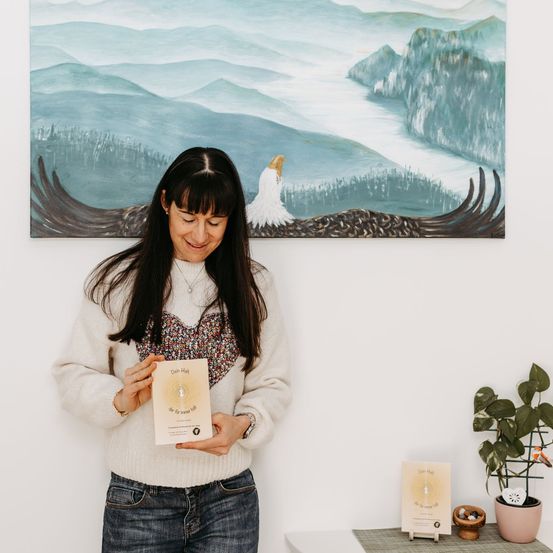 Eine Frau mit langen schwarzen Haaren hält ein Buch mit dem Titel 'Wage es zu träumen' vor einem Gemälde einer friedlichen Landschaft. Sie trägt einen weißen Pullover und Jeans.