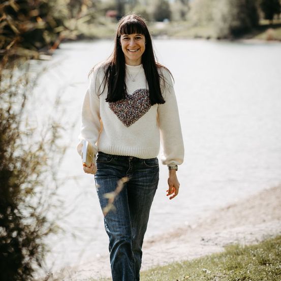Eine Frau mit langen Haaren und einem herzförmigen Pullover geht lächelnd und ein Buch haltend am See entlang.