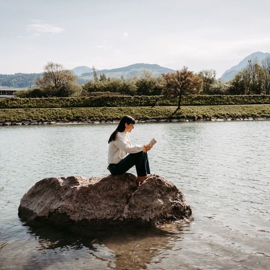 Eine Frau liest ein Buch auf einem Felsen im Fluss, mit Bergen und Grün im Hintergrund.
