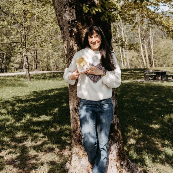 Eine Frau hält ein Buch mit einem Herzmuster auf ihrem Pullover und steht gegen einen Baum in einem Park mit einem Picknicktisch im Hintergrund.