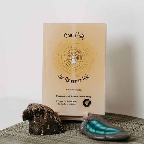 Ein Buch mit dem Titel 'Dein Halt, der für immer halt' steht auf einem Tisch. Es wird von einem kleinen dekorativen Gegenstand in Form einer Eule und einem Schuh mit Federdesign begleitet.