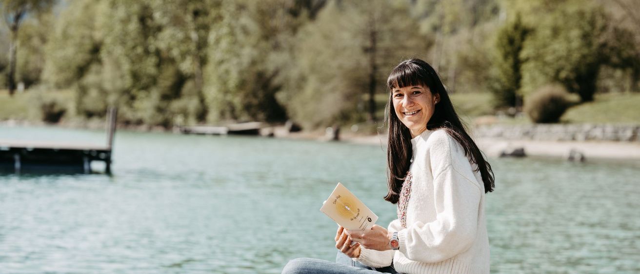 Eine Frau lächelt, hält ein Buch in der Hand und sitzt auf einem Steg mit einem See und Bäumen im Hintergrund.