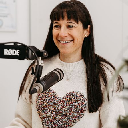 Eine Frau mit langen Haaren und Pony, die ein herzförmiges Sweatshirt trägt, lächelt in die Kamera, während sie in ein Mikrofon mit dem Wort RODE spricht.