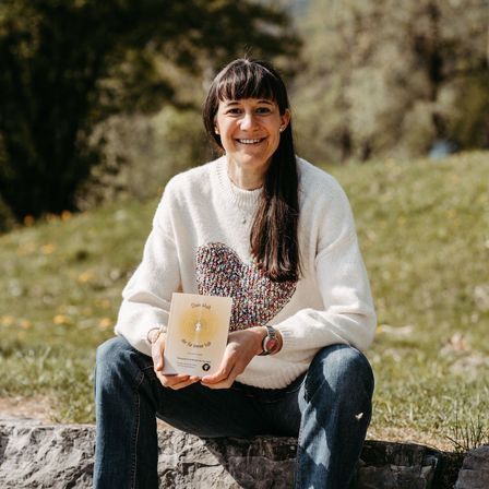 Eine Frau sitzt auf einem Felsen und lächelt, während sie ein Buch in den Händen hält. Sie trägt einen weißen Pullover mit Herzdesign und eine Uhr. Das Buch hat Text darauf. Sie befindet sich in einem Grasbereich mit Bäumen im Hintergrund.