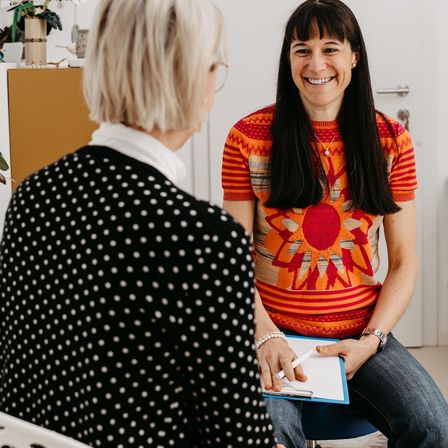 Eine Frau mit einem gepunkteten Oberteil unterhält sich mit einer anderen Frau in einem Sonnenblumen-Pullover, beide sitzen in einem Raum. Die sitzende Frau hält eine Clipboard und einen Stift.