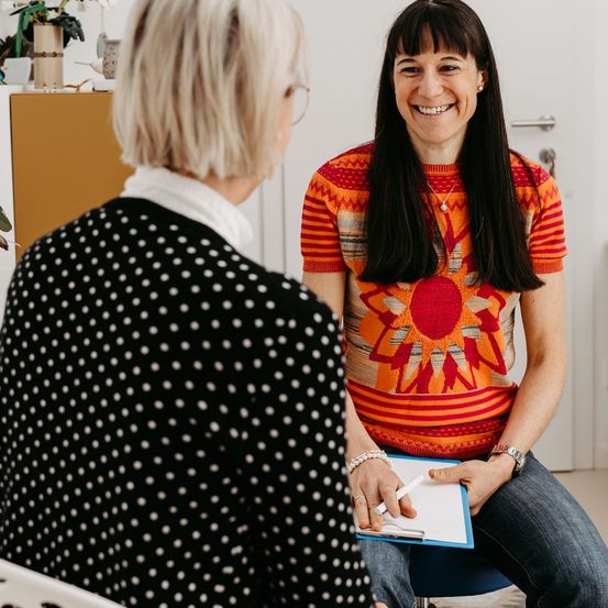Eine Frau mit einem gepunkteten Oberteil unterhält sich mit einer anderen Frau in einem Sonnenblumen-Pullover, beide sitzen in einem Raum. Die sitzende Frau hält eine Clipboard und einen Stift.