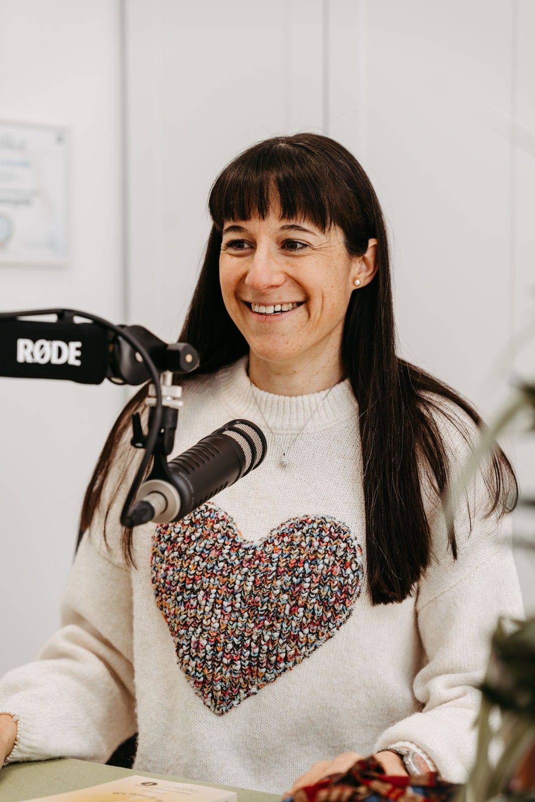 Eine lächelnde Frau steht vor einem Mikrofon und trägt einen weißen Pullover mit einem bunten Herzdesign. Sie scheint sich in einem Aufnahmestudio zu befinden.