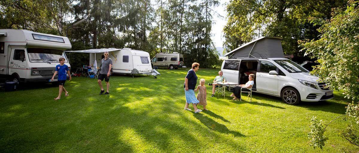 Eine Familie zeltet mit ihren Wohnmobilen auf einem üppigen grünen Feld. Sie sind von Bäumen und Bergen umgeben.
