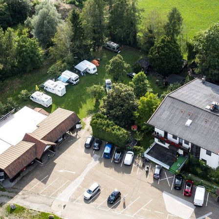 Eine Draufsicht auf einen Campingplatz mit mehreren geparkten Wohnmobilen, einem Gebäude mit braunem Dach und einem Parkplatz mit mehreren Autos und Lieferwagen.