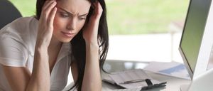 Eine Frau mit den Händen auf dem Kopf sitzt an einem Schreibtisch mit Computer und Papieren und sieht gestresst aus.