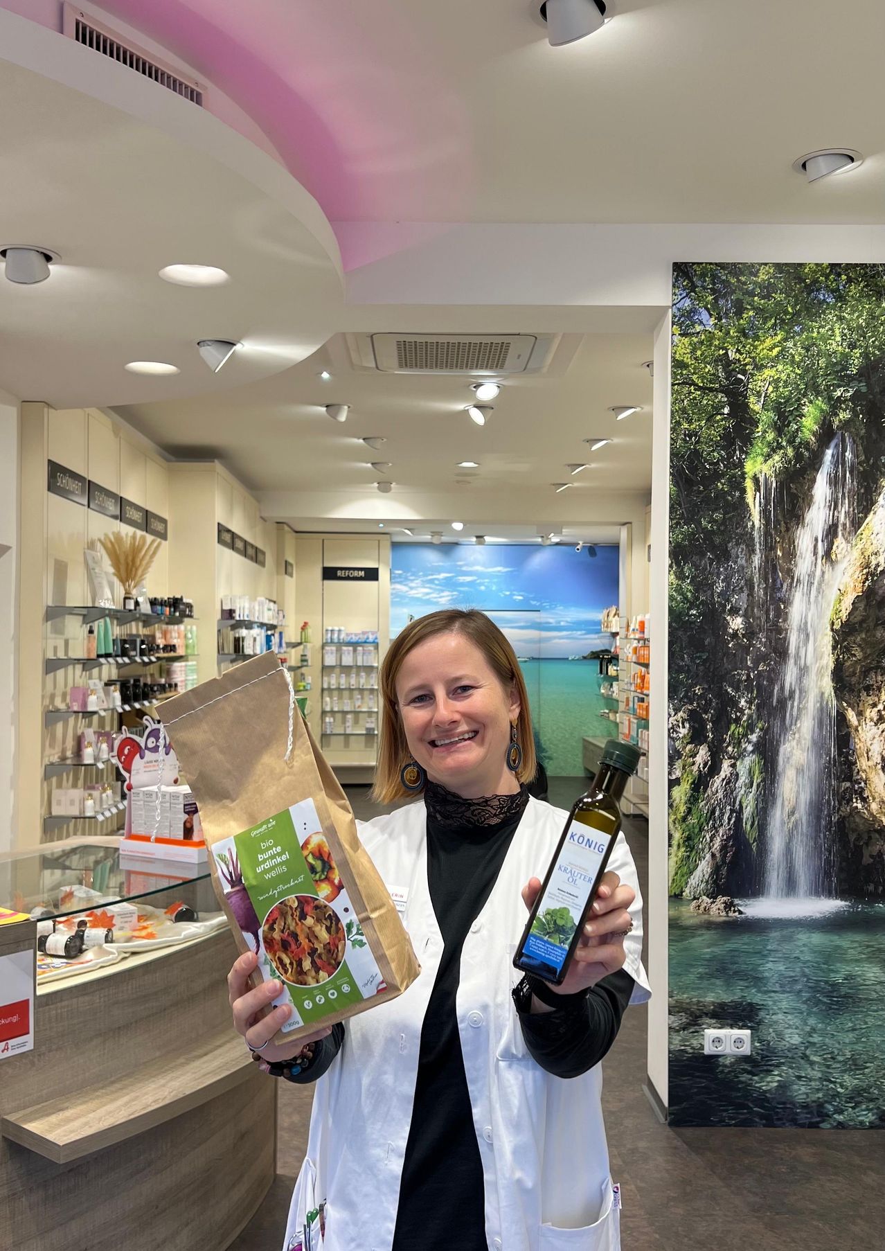 Eine Frau in einem weißen Laborkittel hält einen Beutel mit Bio-Gemüse und eine Flasche Öl in einem Geschäft mit Wasserfalldekoration.