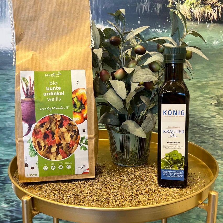 Ein Beutel mit Bio-Kräutermischung, eine Flasche Konig-Kräuteröl und eine Topfpflanze mit Beeren stehen auf einem goldenen Tablett vor einem Wasserfall.