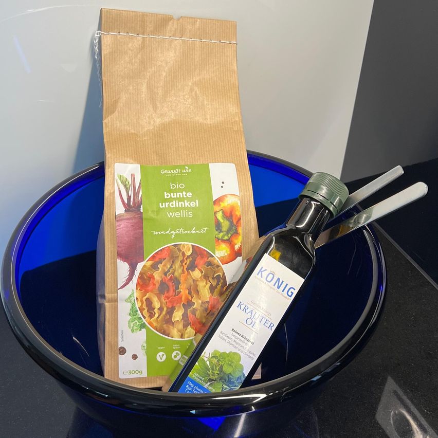 Eine blaue Schüssel mit einer Flasche Olivenöl, einem Beutel mit Bio-Rüben- und Grünzeugmix und einem Messer.