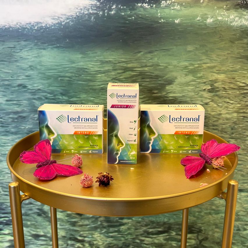 Drei Schachteln Lecternal Allergie-Medikament auf einem goldenen Tablett mit rosa Schmetterlingen und Blumen. Der Hintergrund zeigt einen grünen Ozean.