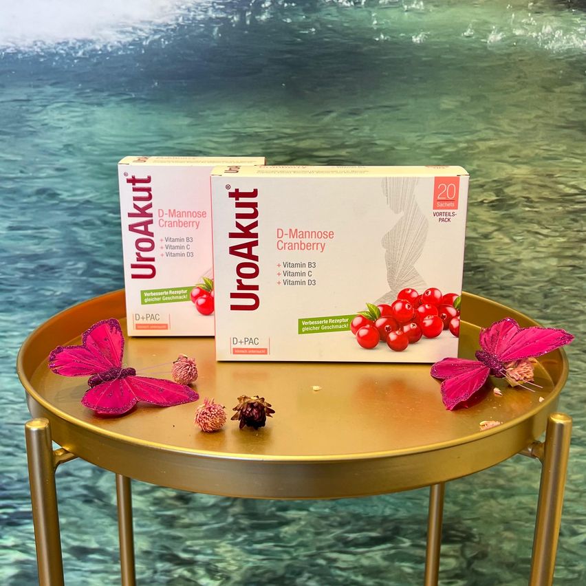 Zwei Schachteln D-Mannose Cranberry stehen auf einem goldenen Tisch mit roten Blumen und Schmetterlingsdesigns. Der Hintergrund ist ein Ozean mit hellgrünem Wasser.