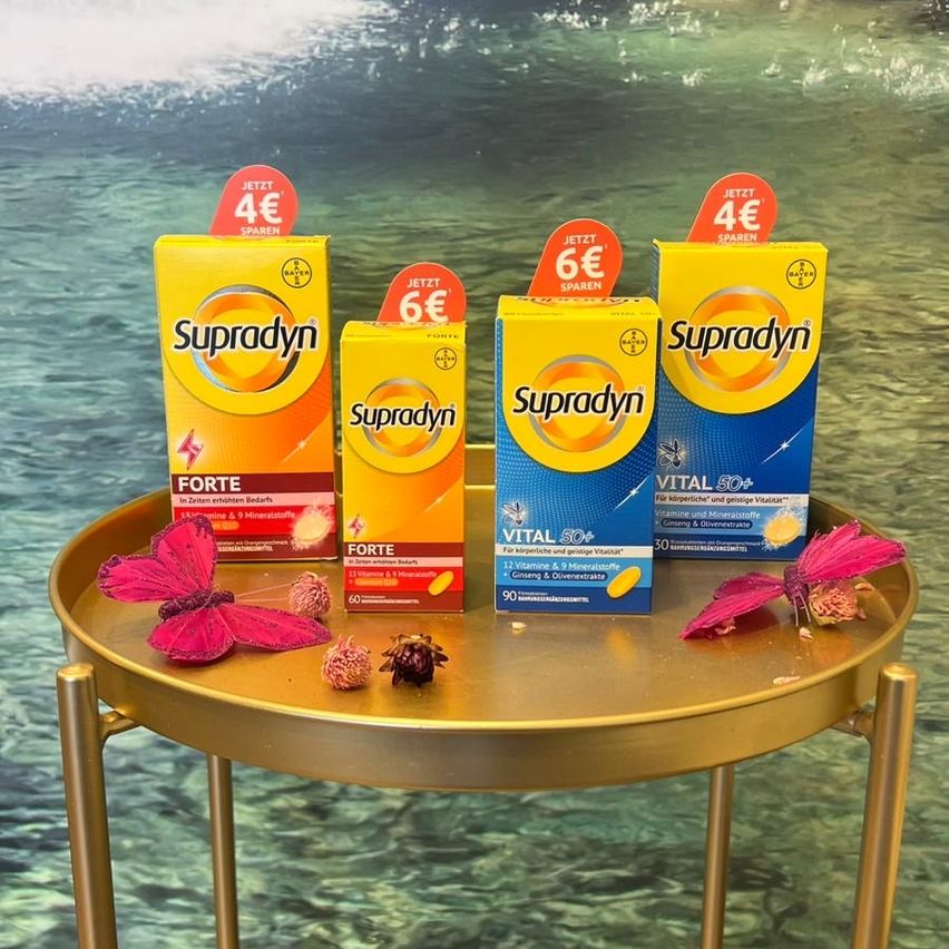 Vier Schachteln Supradyn-Vitamine stehen auf einem goldenen Tisch. Jede Schachtel hat ein anderes Etikett: Forte, Forte, Vital 50+ und Vital 50+. Auf dem Tisch befinden sich zwei rosa Schmetterlinge und einige Blumen.