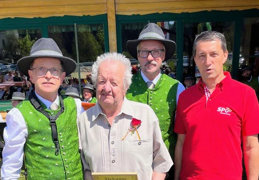 Fünf Männer posieren für ein Foto, einer trägt ein rotes Hemd und eine Blume in der Tasche. Hinter ihnen steht ein Gebäude mit Glasfenstern.