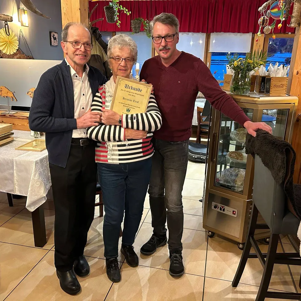 Drei Menschen stehen in einem Restaurant und lächeln. Eine Frau hält ein Zertifikat. Ein Mann neben ihr trägt einen Pullover, während der andere ein weinrotes Hemd trägt. Hinter ihnen steht eine Vitrine mit Essen, und ein Fenster zeigt das Freie.
