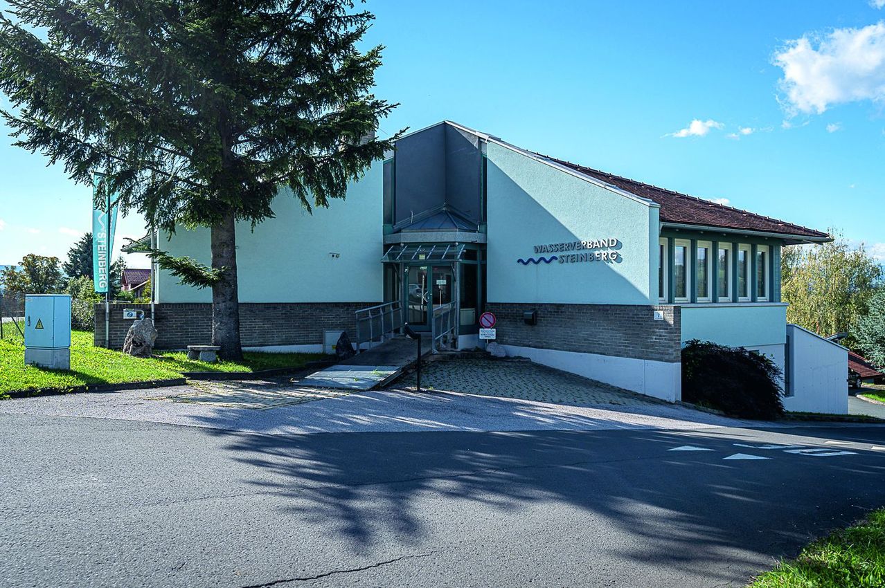 A modern building with a blue wall and a sign reading Wasserverband Steinberg. The entrance is accessible with a ramp.