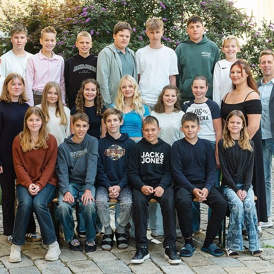 Eine Gruppe von Menschen, wahrscheinlich eine Familie, posiert für ein Foto im Freien. Sie stehen und sitzen auf einer Bank vor einem Gebäude mit Pflanzen.