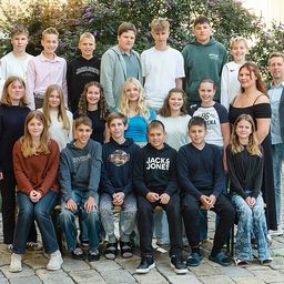 Eine Gruppe von Menschen, wahrscheinlich eine Familie, posiert für ein Foto im Freien. Sie stehen und sitzen auf einer Bank vor einem Gebäude mit Pflanzen.