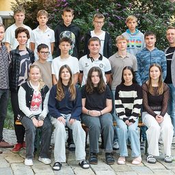 Eine Gruppe von Schülern und Lehrern posiert für ein Foto im Freien, mit zwei Lehrern links und rechts, umgeben von Grün.