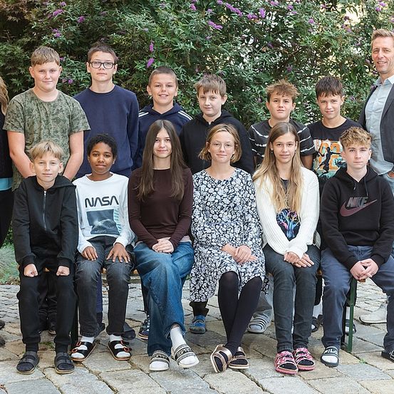 Eine Gruppe von Kindern und Erwachsenen posiert für ein Foto im Freien. Die Kinder sitzen auf einer Bank, während die Erwachsenen hinter ihnen stehen. Im Hintergrund befinden sich grüne Bäume mit lila Blüten.