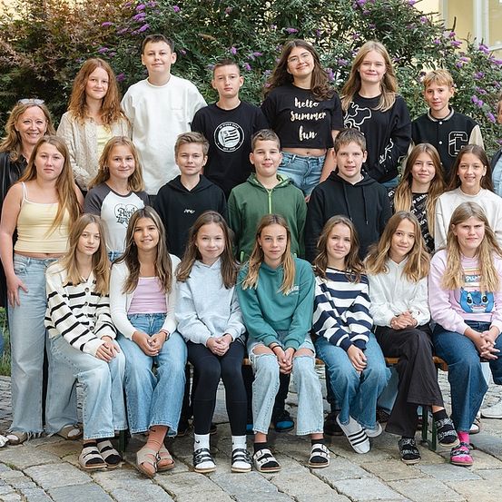 Eine Gruppe von Schülern und Lehrern posiert für ein Foto vor einem Gebäude mit Pflanzen und Fenstern. Einige tragen Brillen, Turnschuhe und Freizeitkleidung.
