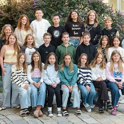 Eine Gruppe von Schülern und Lehrern posiert für ein Foto vor einem Gebäude mit Pflanzen und Fenstern. Einige tragen Brillen, Turnschuhe und Freizeitkleidung.