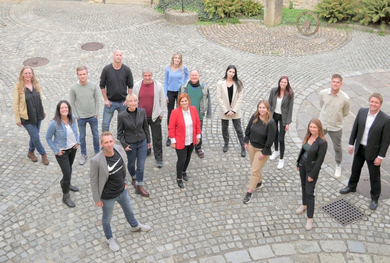Eine Gruppe von Menschen in verschiedenen Kleidungsstücken steht auf einer Kopfsteinpflasterstraße und lächelt für ein Foto. Hinter ihnen sind Pflanzen und ein Steinrad.
