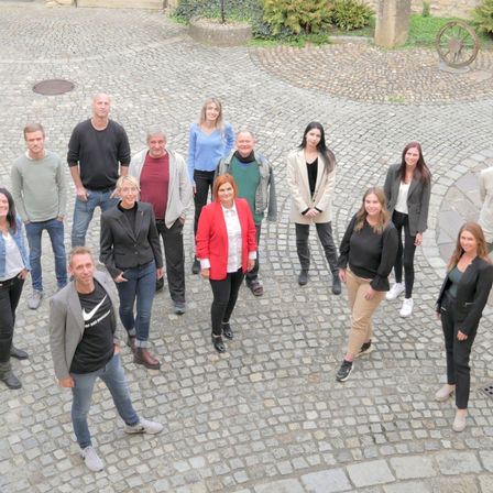 Eine Gruppe von Menschen in verschiedenen Kleidungsstücken steht auf einer Kopfsteinpflasterstraße und lächelt für ein Foto. Hinter ihnen sind Pflanzen und ein Steinrad.