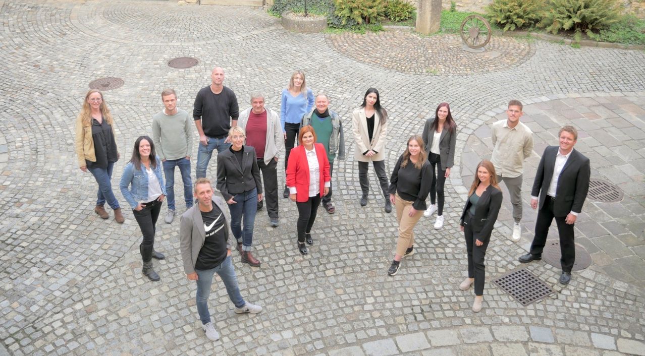 Eine Gruppe von Menschen in verschiedenen Kleidungsstücken steht auf einer Kopfsteinpflasterstraße und lächelt für ein Foto. Hinter ihnen sind Pflanzen und ein Steinrad.