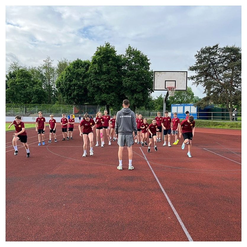 Eine Gruppe von Schülern in roten Uniformen läuft auf einer Laufbahn, angeführt von einem Mann in Grau. Ein Basketballkorb befindet sich hinter ihnen.